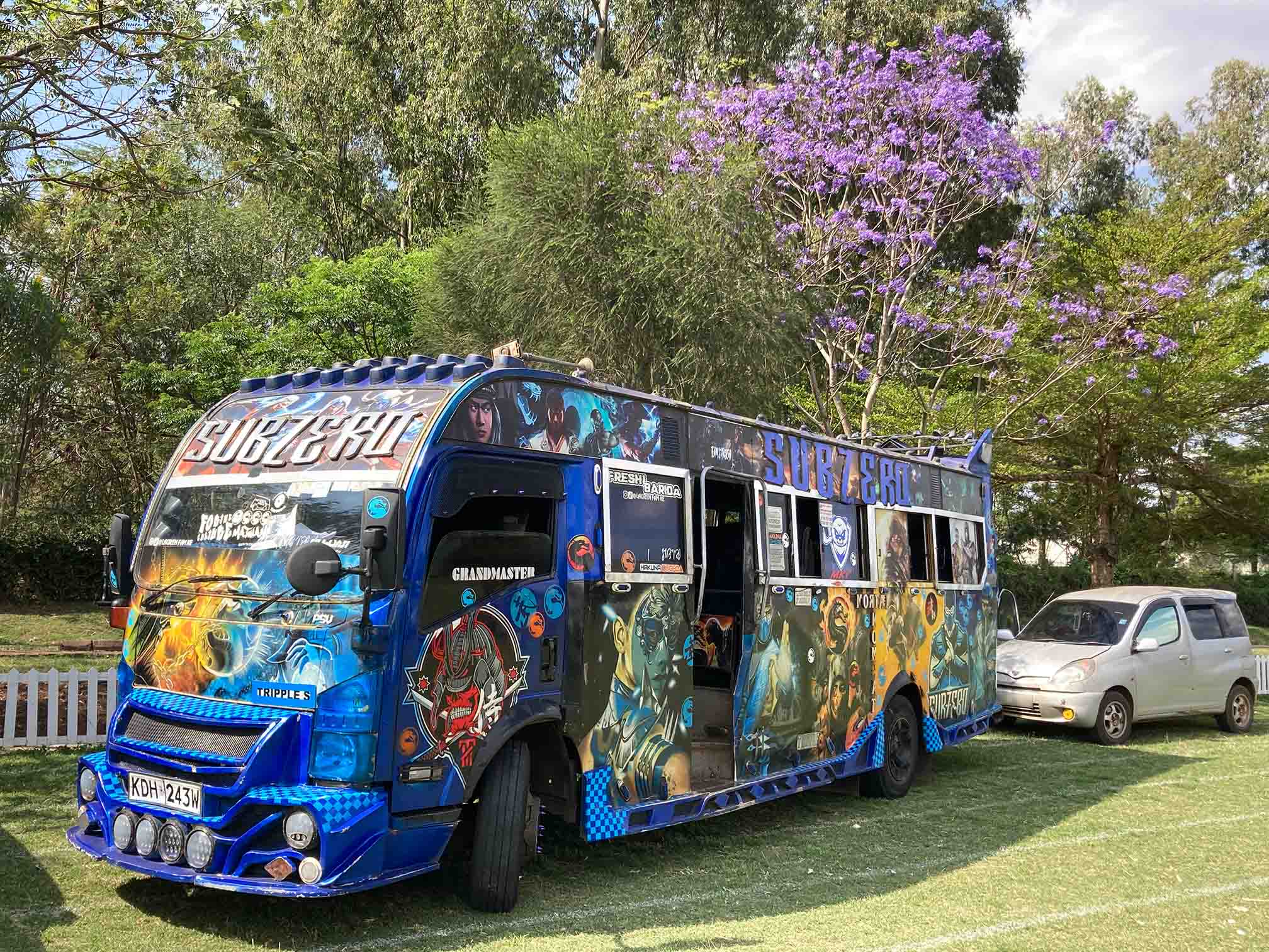 A blue Kenyan matatu (minibus) named “SUBZERO,” featuring bold graffiti art inspired by video game characters and fantasy themes, parked on a grassy area next to a small car.