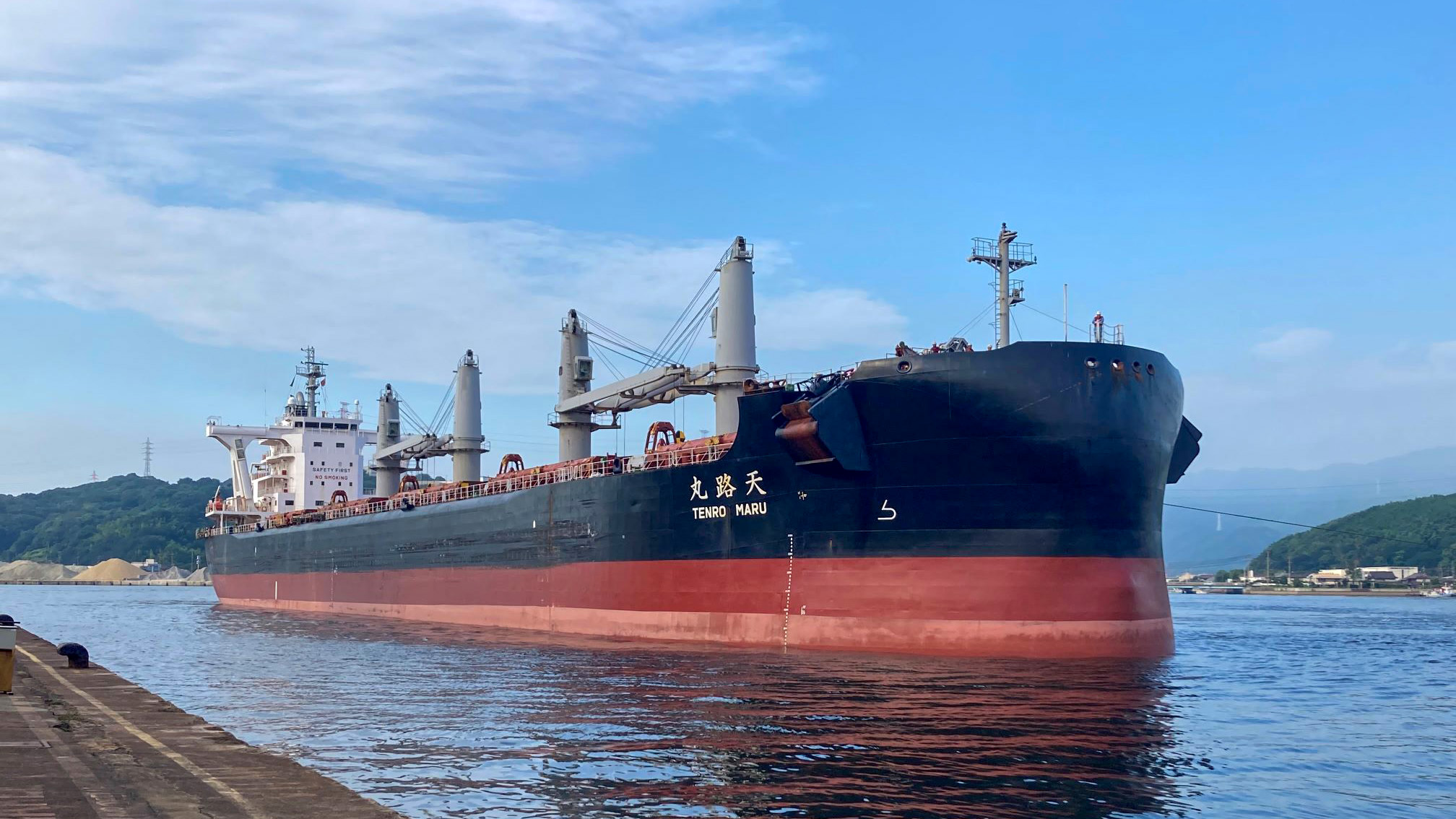 The dry bulk carrier ‘TENRO MARU’ is docked at a port, with its large hull and cranes visible against a backdrop of hills and a clear sky, ready for cargo operations.