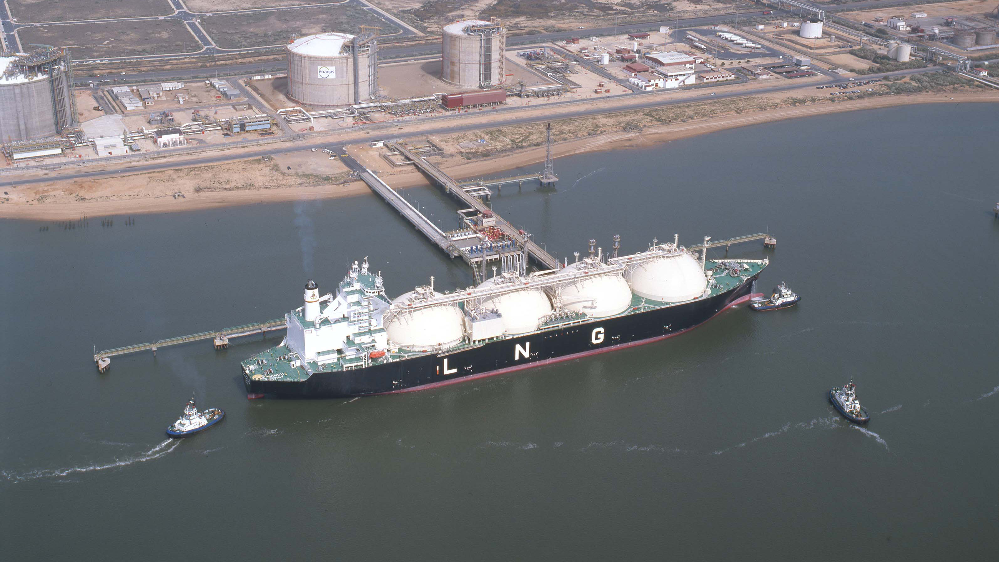 An LNG tanker is docked at a terminal with storage tanks in the background, connected by pipelines, showcasing the infrastructure and operations involved in LNG transport.