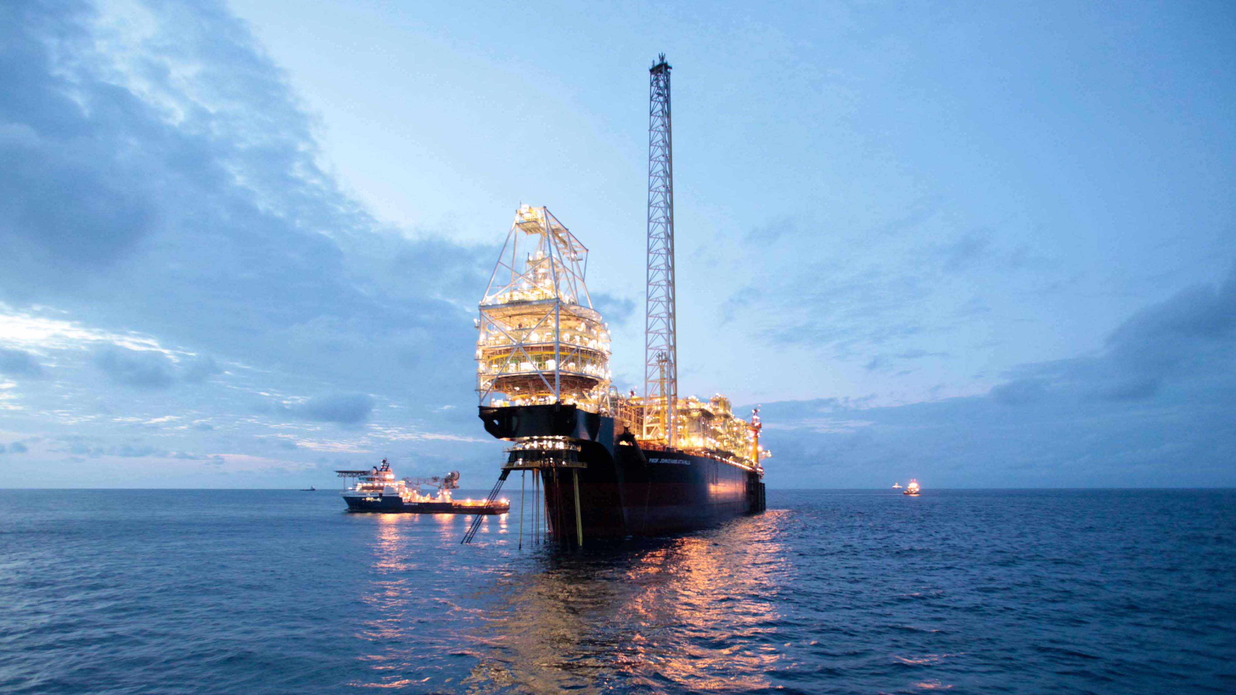 An illuminated offshore oil platform with support vessels nearby, sitting in calm waters during twilight, showcasing the scale of offshore energy operations.