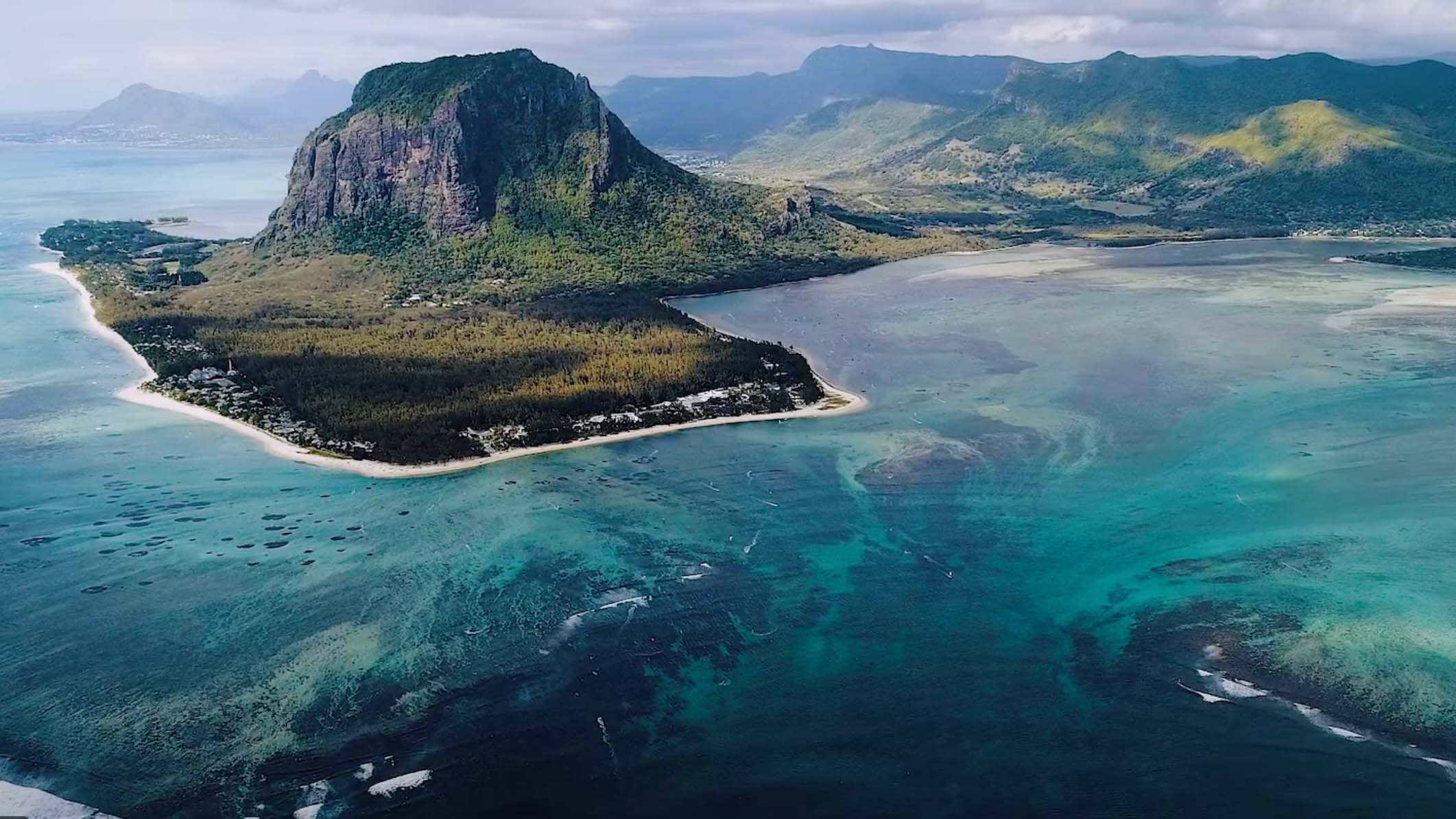 An aerial view of Mauritius, featuring a towering mountain surrounded by lush greenery, white sandy beaches, and clear turquoise waters, highlighting the island’s natural beauty.