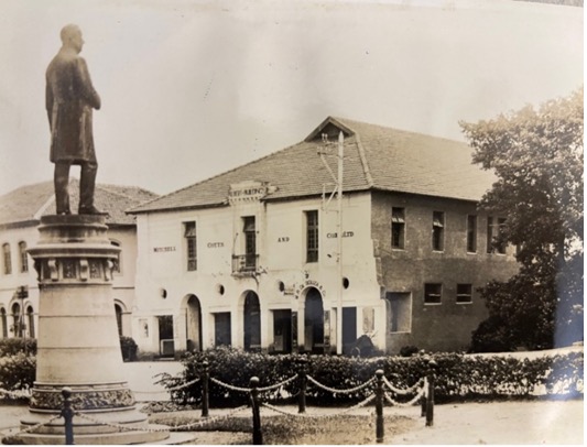 A black-and-white photograph of the pre-war Mombasa Resident Office (1929-1941), a colonial-style building with “MITCHELL COTTS AND CO LTD” written on its facade, and a statue of a man in the foreground.