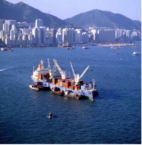 Semi-con ship: Atorantikkumaru in a bustling harbor with skyscrapers and hills in the background, equipped with cranes and docked alongside smaller vessels.