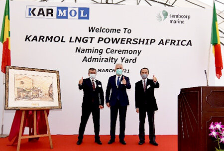 Three men in suits and masks give a thumbs-up during the naming ceremony of ‘KARMOL LNGT POWERSHIP AFRICA’ at Admiralty Yard, with a framed artwork and flags in the background.