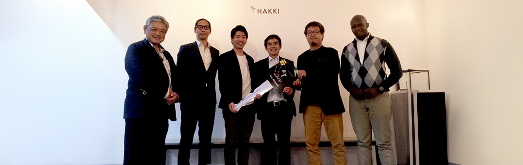 Six men stand together in front of a wall with the logo ‘HAKKI,’ symbolizing a partnership between Kilimol, Karasawa, and Hakki, with representatives from each company present.