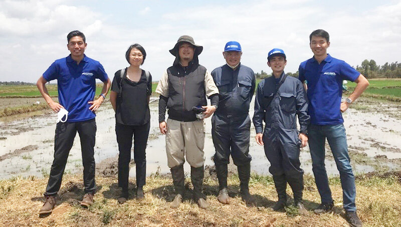 A group of five people, including representatives in blue shirts and a man in farming attire, stand together in a field as part of the MOL and Kilimol Double Feather MOU, symbolizing agricultural collaboration.