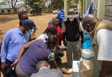 A group of people, including local workers and a representative in a hat and vest, gather around equipment as part of the MOL and Karasawa Rice Mills subsidy project, supporting agricultural initiatives.