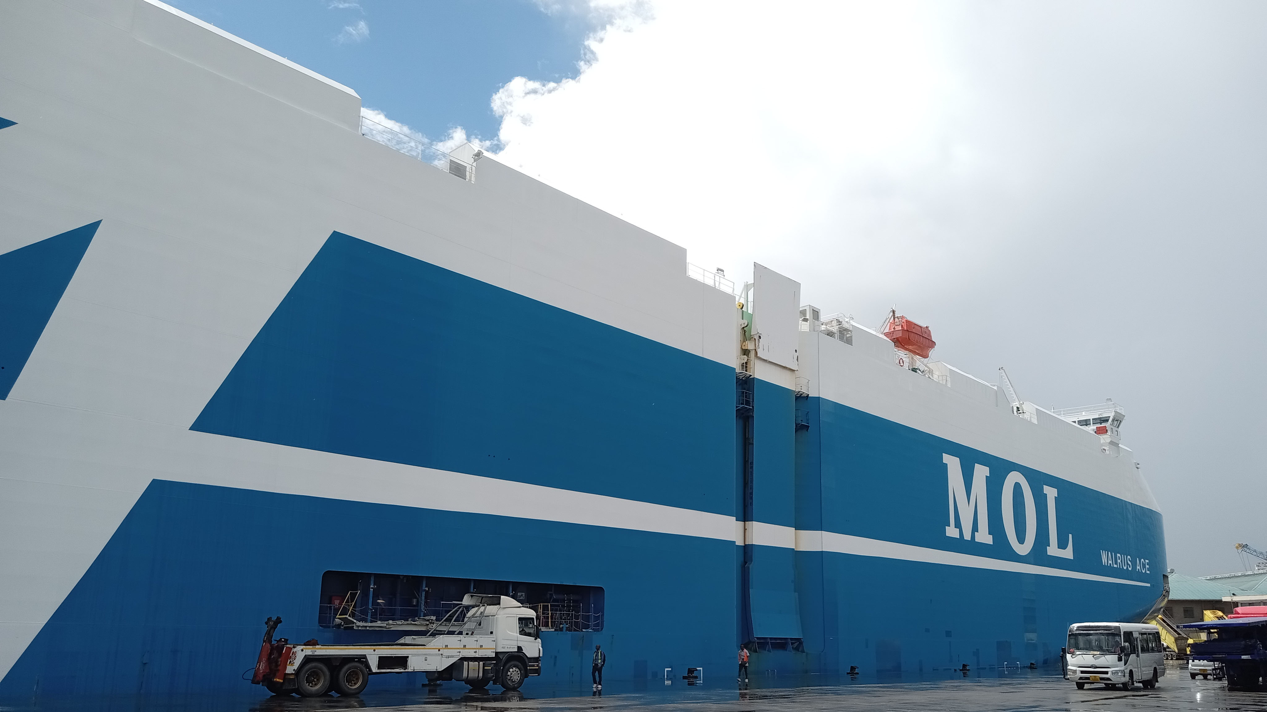 The MOL car carrier ship ‘WALRUS ACE’ is docked at Dar es Salaam port, with a truck and people in the foreground, showcasing the scale of product transport operations.