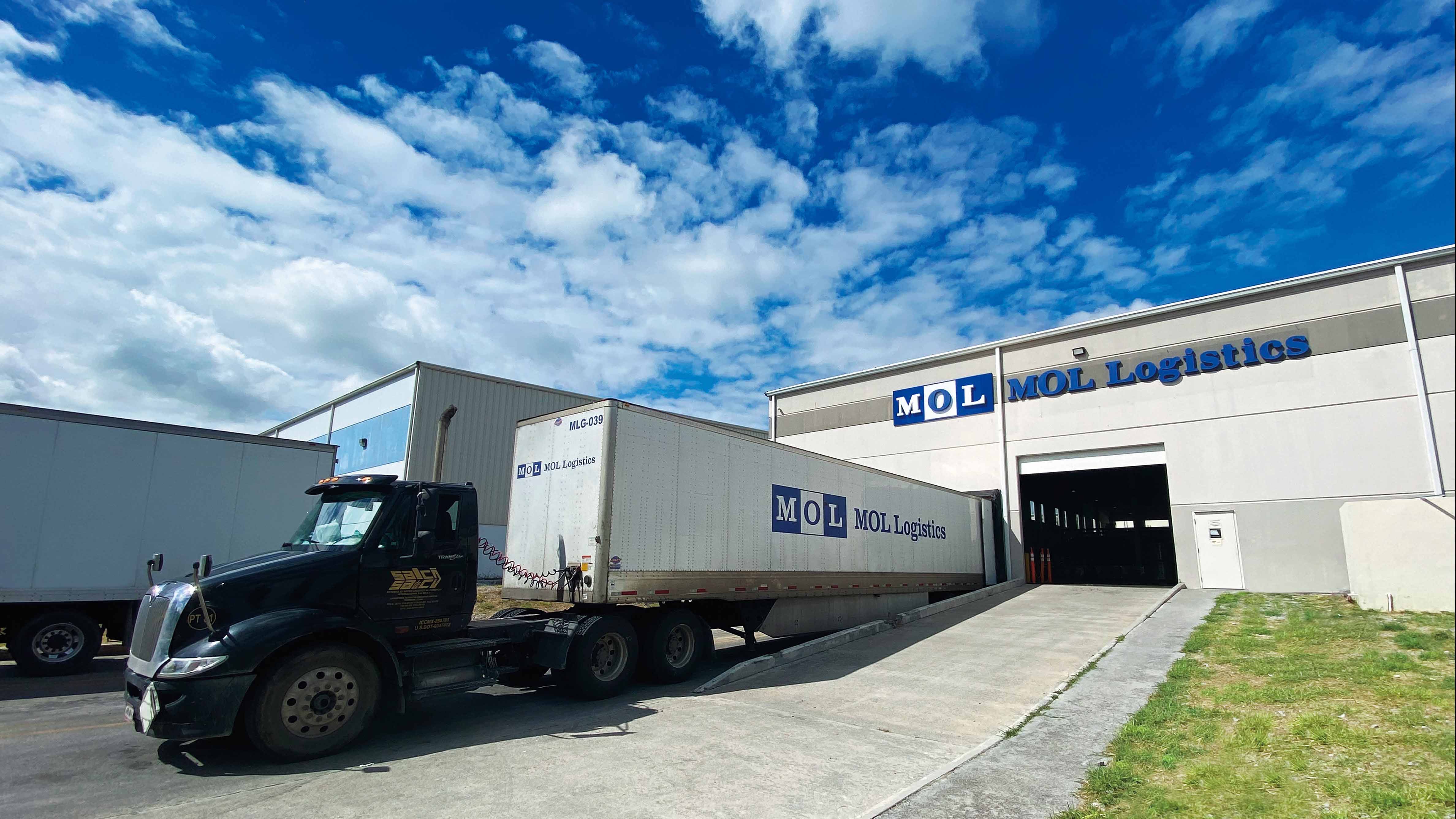 A black truck is parked in front of the MOL Logistics warehouse, with a large trailer labeled ‘MOL Logistics’ being loaded, illustrating transportation and logistics operations under a clear sky.